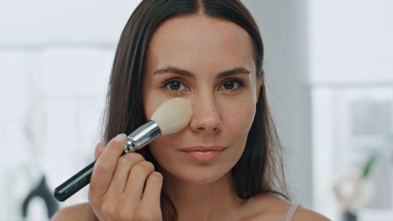 Žena nanosi puder četkicom na lice ispred ogledala, fokus na kontakt četkice sa kožom obraza